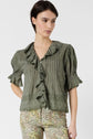 Woman wearing an olive-green, ruffled blouse with short sleeves and floral-patterned pants, standing against a plain white background.