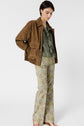 Woman wearing a brown suede jacket, olive green ruffled blouse, and floral patterned flared pants stands barefoot against a plain white background, looking downward with hands in her jacket pockets.