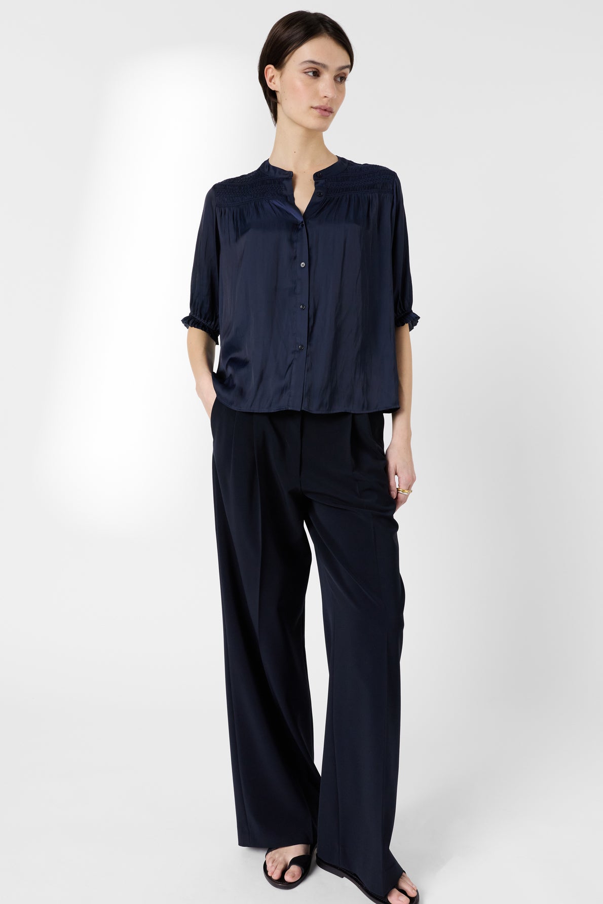 Person standing against a plain light background, wearing a loose navy blouse with buttons, wide-leg matching navy trousers, and black sandals, with hands in pockets and a neutral expression.