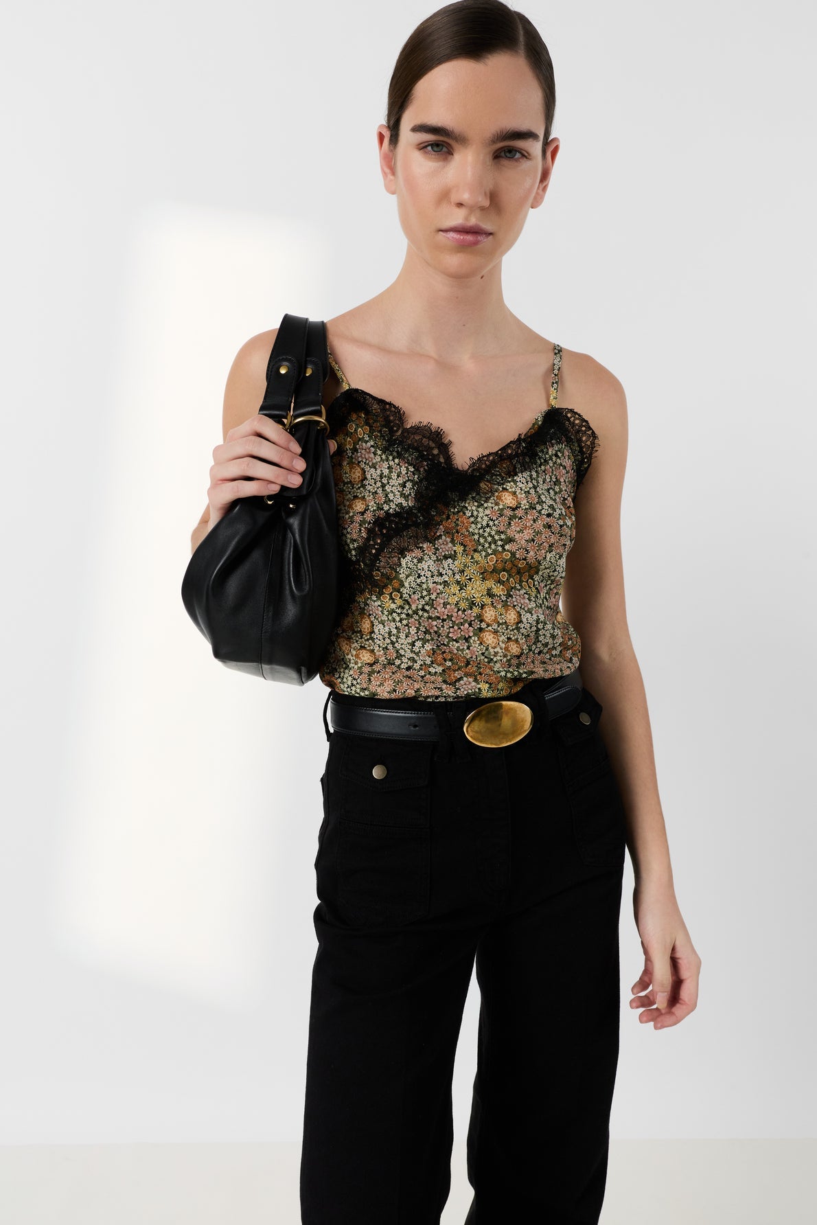 A woman wearing a floral lace-trimmed camisole, black high-waisted pants with a large gold belt buckle, and holding a black shoulder bag, stands against a plain white background.