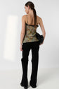 A person with long brown hair in a low ponytail wears a floral-patterned sleeveless top, black pants, and holds a black bag, standing against a white background facing away.