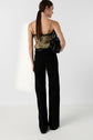 Woman standing in a studio, facing away. She wears a floral tank top, black high-waisted pants, and holds a black handbag. Her brown hair is tied in a low ponytail. The background is plain white.