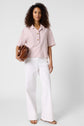 A person with curly hair wearing a chemise rayée à manches courtes et coupe ample, in light pink stripes, paired with white wide-leg pants, holding a brown handbag and wearing sandals, stands against a plain white background.