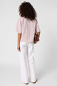 Person standing facing away, wearing a light pink chemise rayée à manches courtes and white pants, holding a brown leather bag by their side against a plain white background.