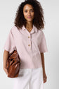 Woman with curly hair wearing a chemise rayée rose et blanche à manches courtes and white pants, holding a large brown leather bag, standing against a plain light background.