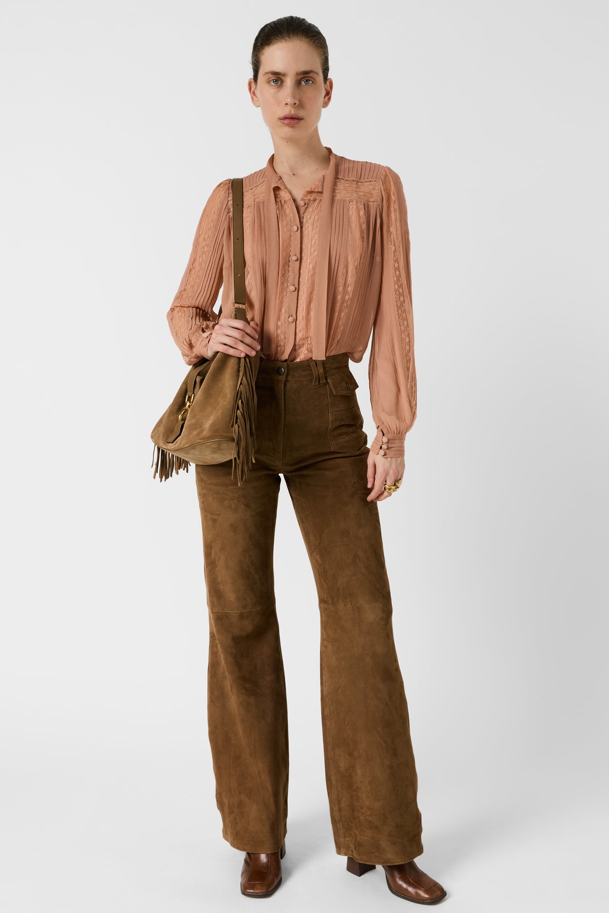 Woman wearing a light pink chemise lavallière, brown high-waisted suede flared pants, brown boots, and carrying a brown fringe bag, standing against a plain white background.