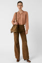Woman wearing a light pink chemise lavallière, brown high-waisted suede flared pants, brown boots, and carrying a brown fringe bag, standing against a plain white background.
