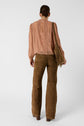 A person with brown hair in a low bun wears a pink chemise lavallière, brown suede pants, and brown shoes, carrying a brown fringed shoulder bag, standing against a plain white background.