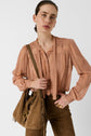 Woman wearing a peach chemise lavallière long-sleeve blouse and brown pants, holding a brown suede fringe bag over her shoulder, standing against a plain white background.
