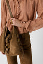 A person wearing a sheer, pleated, dusty pink chemise lavallière and brown suede pants holds a brown suede handbag with a gold buckle and fringe detail. Only the torso and handbag are visible.