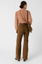 A woman with her hair in a low bun wears a sheer pink chemise lavallière, high-waisted brown suede pants, heeled boots, and a brown fringed shoulder bag, standing with her back to the camera against a white background.
