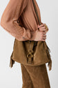 A person in a peach chemise en dentelle blouse and brown suede pants holds a large brown suede shoulder bag with gold hardware and fringe details, suggesting an air of édition limitée luxury. Only the torso and hands are visible.