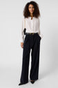A woman with curly hair wears a white button-up blouse, black wide-leg trousers, black pointed shoes, and a black shoulder bag with a gold chain strap, standing against a plain background.