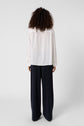 A person with curly hair stands facing away, wearing a loose white blouse and wide-leg black pants against a plain light background.