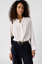 A person with curly hair wears a white button-up blouse and black pants with a belt, carrying a black shoulder bag with a gold chain, standing against a plain light background.