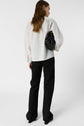 A person with long brown hair, wearing a white chemise Calixte with col mao and black pants, stands facing away while holding a black handbag under their left arm against a plain white background.