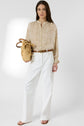 Woman standing against a white background, wearing a beige chemise manches bouffantes with delicate patterns, white high-waisted pants, brown belt, and holding a woven straw bag. She has long brown hair and is wearing sandals.