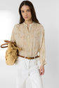Woman with long brown hair wearing a beige chemise manches bouffantes patterned blouse and white pants, holding a woven straw bag. She has a neutral expression and stands against a plain light background.