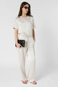 A woman in sunglasses wears a cream-colored t-shirt and matching wide-leg pants. She holds a black clutch with a gold chain and wears sandals, posing against a plain white background.