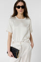 Woman with long brown hair wearing black sunglasses, an off-white short-sleeve shirt, and matching pants, holding a black clutch with a gold chain, standing against a plain white background.