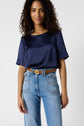 Woman with curly hair wears a navy short-sleeve blouse tucked into high-waisted blue jeans with a brown belt, standing against a plain light background.