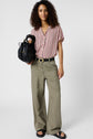 Woman with curly hair wearing a striped short-sleeve shirt, olive wide-leg pants, black belt, and sandals, holding a black handbag, stands against a plain white background.