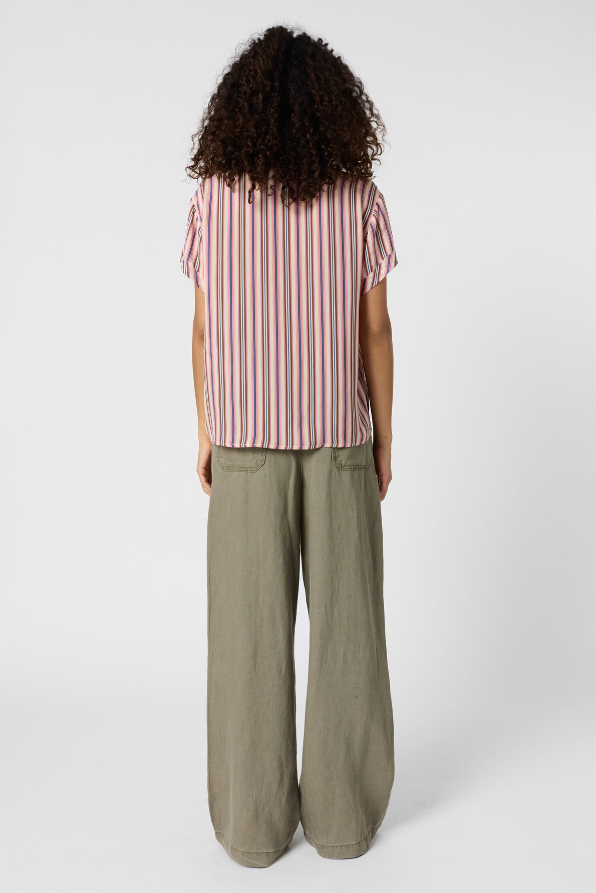 Person standing facing away, wearing a short-sleeve, vertically striped pink and white shirt, and loose-fitting, light olive green pants, against a plain white background. Their curly hair reaches their shoulders.