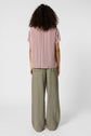 Person standing facing away, wearing a short-sleeve, vertically striped pink and white shirt, and loose-fitting, light olive green pants, against a plain white background. Their curly hair reaches their shoulders.