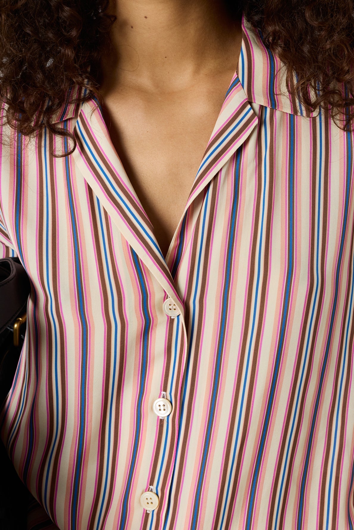 A close-up of a person wearing a collared shirt with vertical pink, blue, black, and white stripes. The shirt is buttoned, and the persons curly hair is visible at the top.