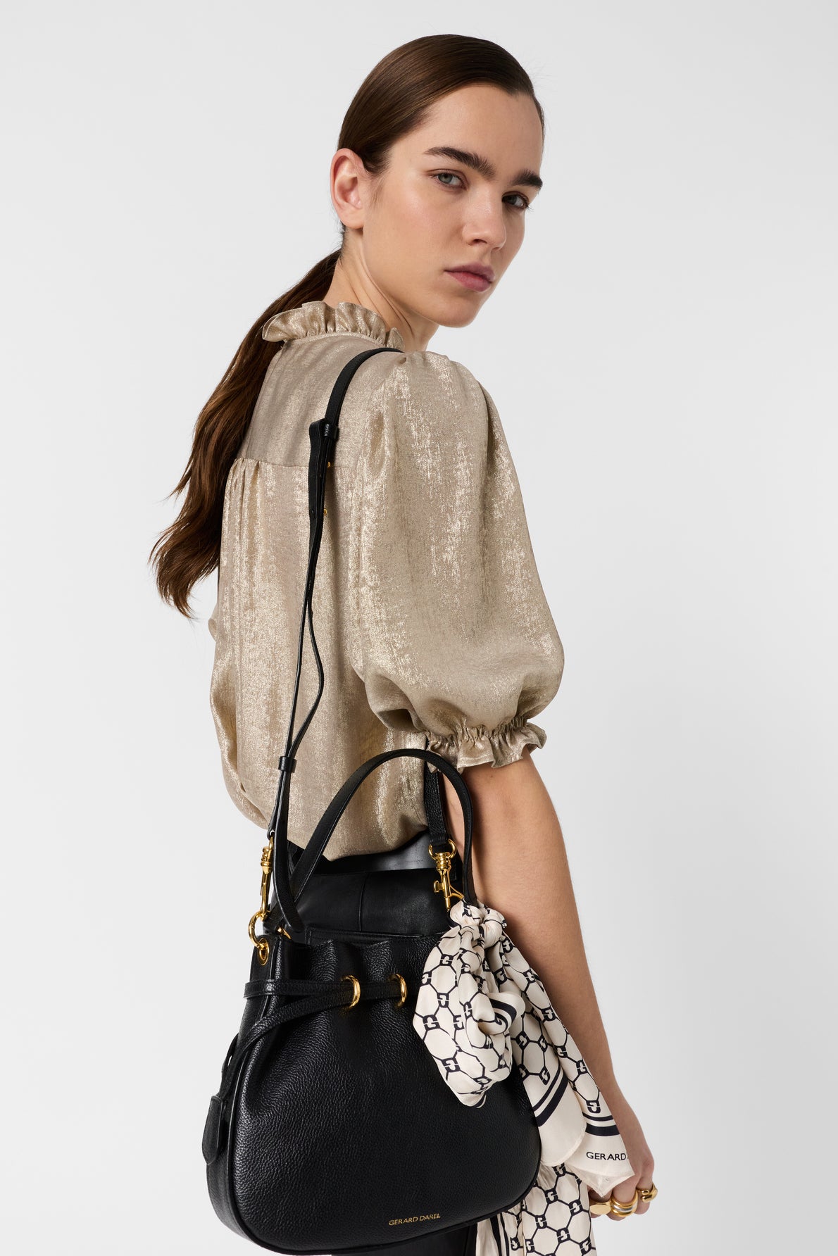Woman with long brown hair, wearing a shiny beige blouse, holds a black handbag with gold hardware. A patterned scarf is tied to the bags handle. She looks over her shoulder against a plain white background.
