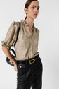 A person wearing a shiny gold blouse with ruffled collar and puffed sleeves, tucked into black belted pants with large pockets, holding a black handbag, standing against a plain white background.