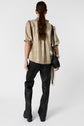 Woman standing with her back to the camera, wearing a shiny gold blouse, black leather pants, and black shoes, carrying a black handbag and a patterned scarf. Her brown hair is tied back in a low ponytail.