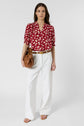 Woman standing against a plain background, wearing a chemise fleurs with manches bouffantes, white wide-leg pants, tan sandals, and carrying a brown handbag.