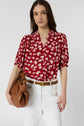 A woman wearing a red chemise fleurs with manches bouffantes and white pants holds a brown handbag and looks at the camera. She has long brown hair and a neutral expression.