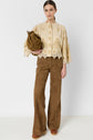 Woman standing against a white wall, wearing a beige embroidered blouse with lace trim crafted from coton seersucker, brown suede wide-leg pants, brown clogs, and holding a brown suede bag. This look is part of an édition limitée collection.