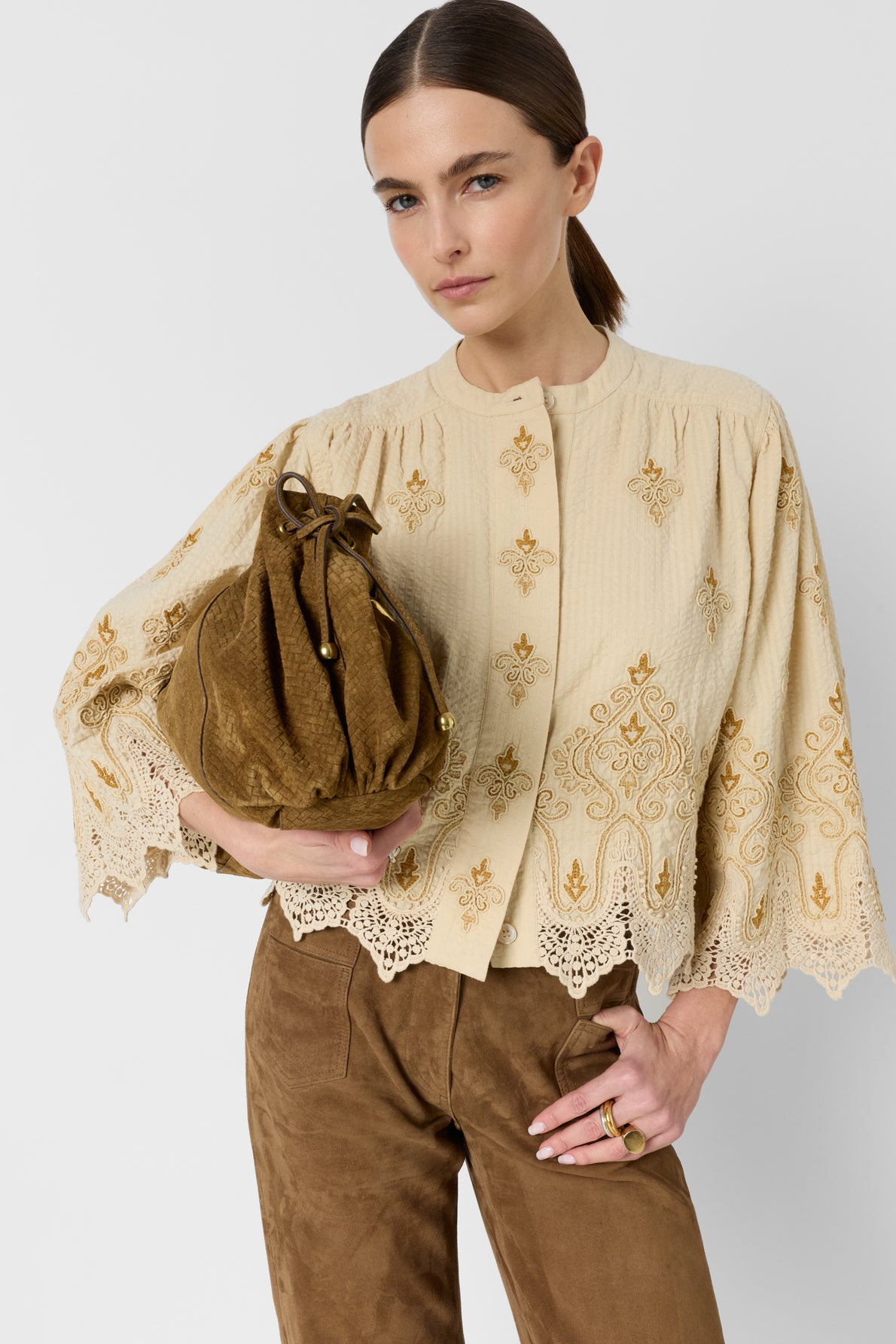 Woman with straight brown hair in a beige chemise épaisse embroidered top and brown pants holds a brown suede drawstring bag, looking confidently at the camera. The outfit, crafted in édition limitée, features lace details and gold accents.