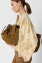 Woman in a cream embroidered chemise épaisse holding a large brown textured bag under her arm, shown from the side against a plain light background.