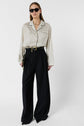 Woman wearing black sunglasses, a chemise satinée à pois with a col tailleur, and high-waisted, wide-leg black pants. She holds a black clutch and stands against a plain white background.