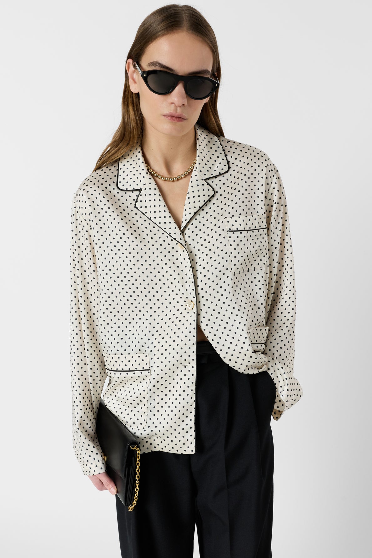 A person wearing black sunglasses, a gold chain necklace, and a white pois chemise satinée button-up shirt with black trousers, holding a black clutch with a gold chain, poses against a plain white background.