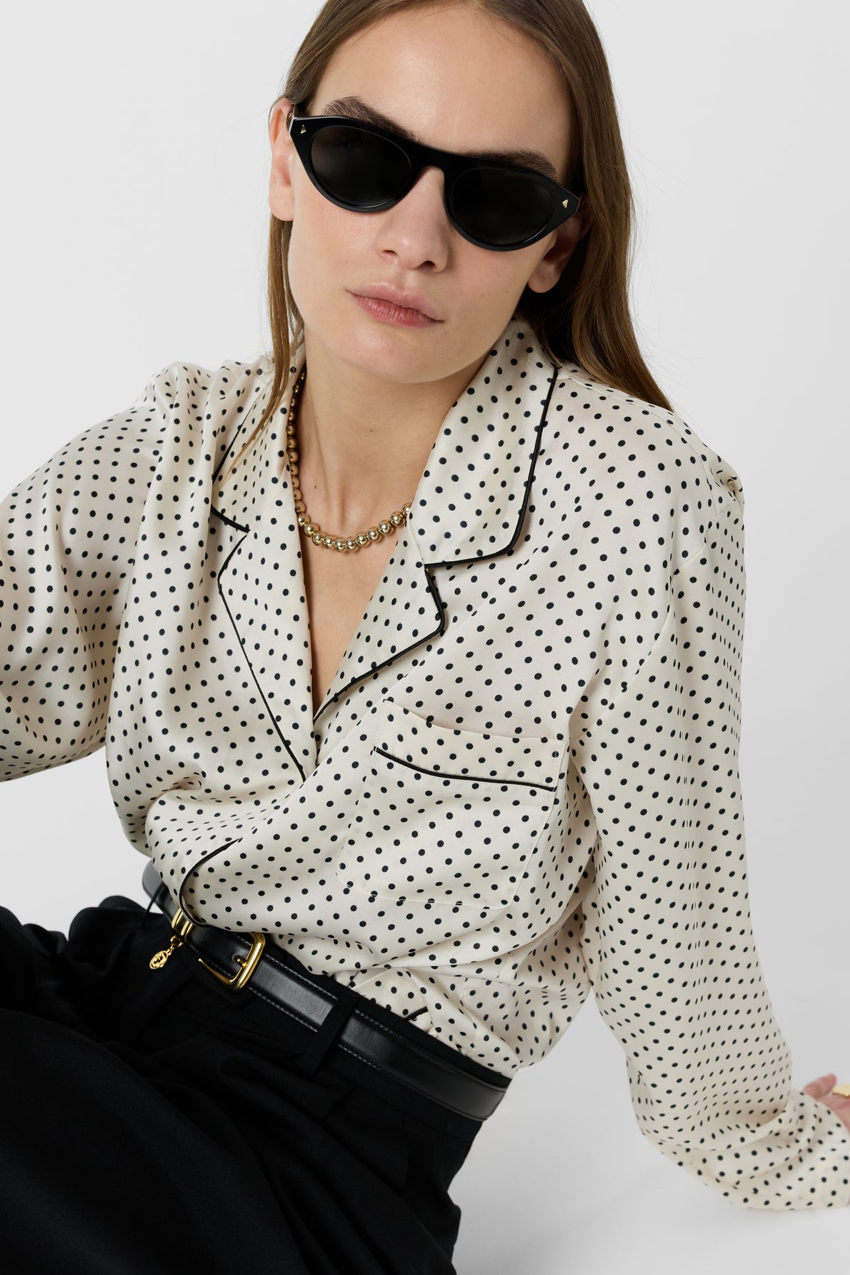 Woman wearing black sunglasses, a gold chain necklace, a cream-colored chemise satinée à pois and black high-waisted pants with a belt, sitting against a plain white background.