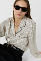 Woman wearing black sunglasses, a gold chain necklace, a cream-colored chemise satinée à pois and black high-waisted pants with a belt, sitting against a plain white background.