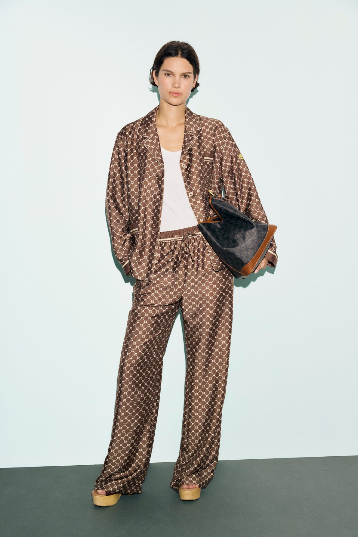 A person stands against a light background, wearing a loose brown patterned pajama-style suit layered over a white tank top, beige platform shoes, and holding a large black geometric handbag. The look is elevated by the addition of an exclusive chemise satinée adorned with an édition limitée monogramme GD.
