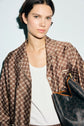 Woman with short dark hair wears a chemise satinée in a brown pattern over a white tank top, holding an édition limitée black patterned handbag on her shoulder, standing against a light background.