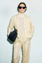 Person wearing a light yellow chemise satin jaune lounge set with a white turtleneck and black sunglasses, holding a black handbag, standing against a plain light background.