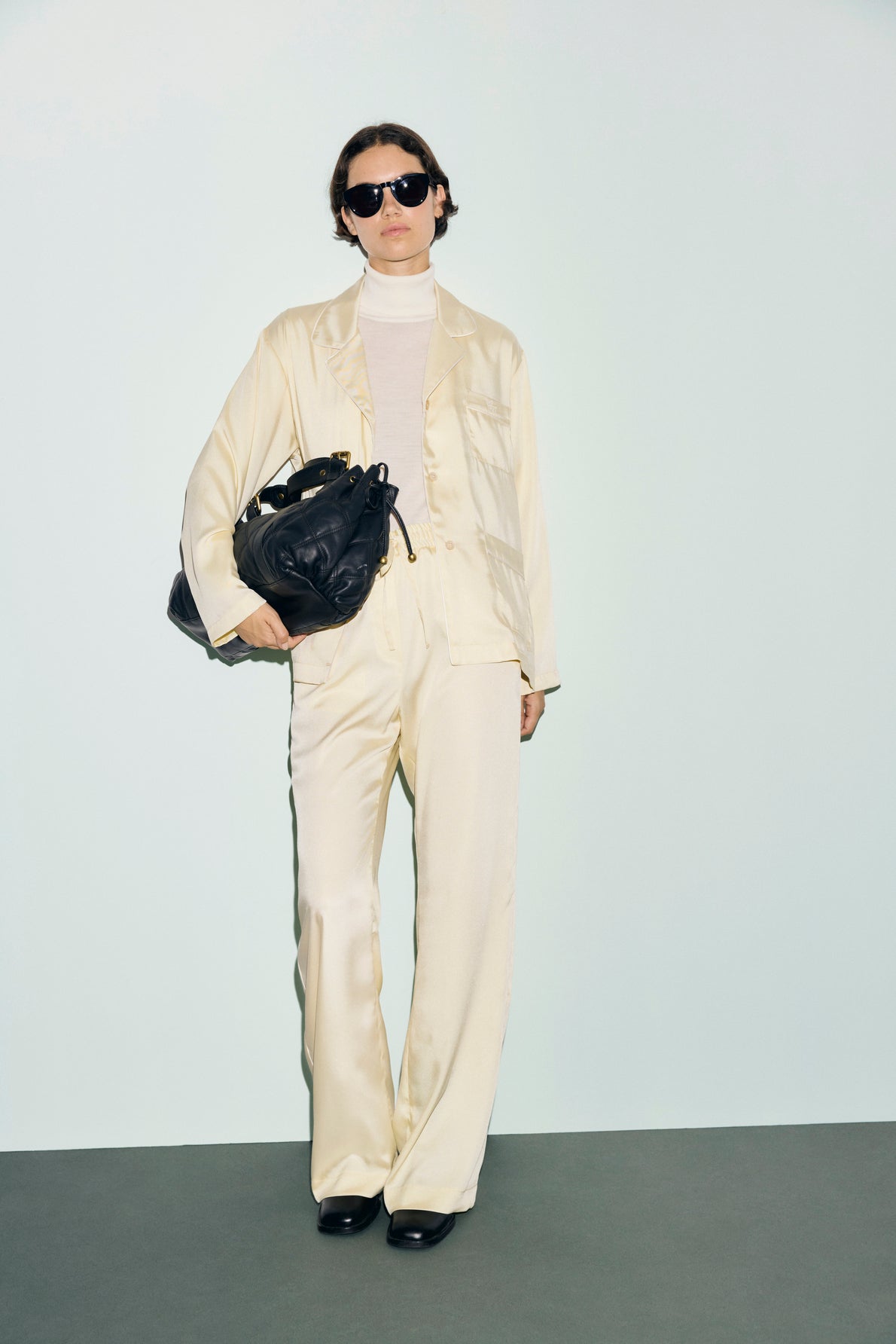 Person wearing a cream-colored satin suit with a chemise satin jaune, white turtleneck, and black sunglasses, standing against a light background, holding a large black bag and wearing black shoes.