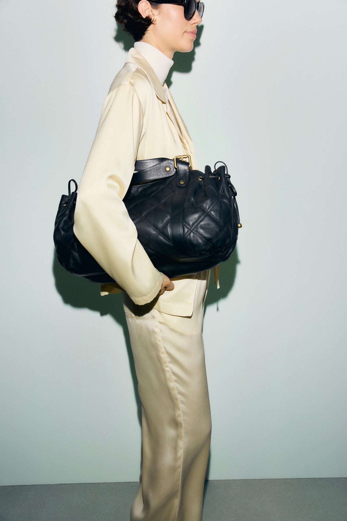 A person in a cream-colored suit and sunglasses, wearing a chemise col tailleur, holds a large, black quilted handbag under their arm against a plain light background.