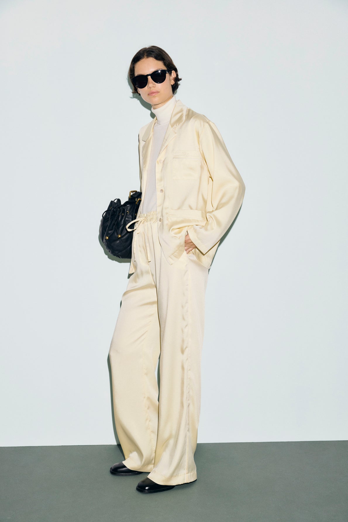 Person wearing sunglasses, a cream-colored satin suit with wide-leg pants, a light turtleneck, and black shoes, holding a black handbag, stands against a plain light background—her look is elevated by a subtle nod to classic tailoring with a chemise col tailleur.