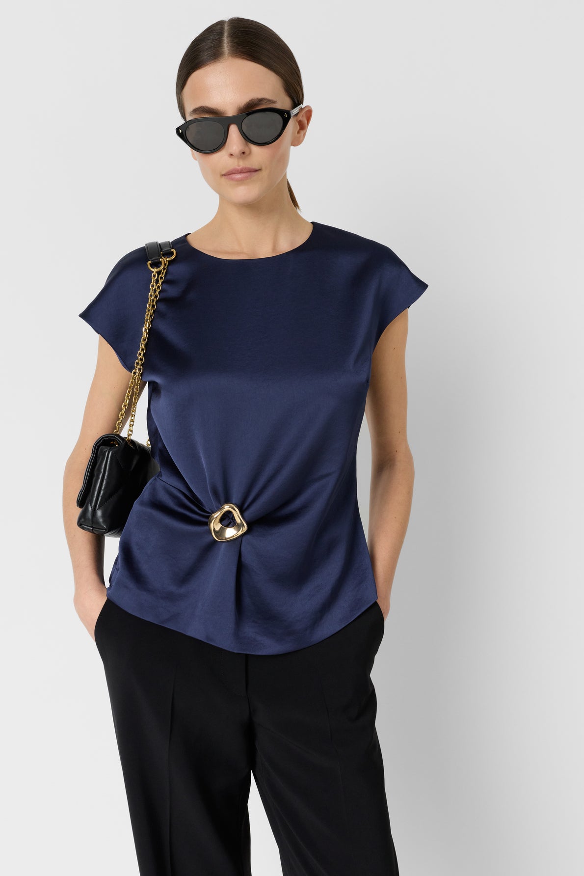 A woman wearing black sunglasses, a satin bleu marine top sans manche with a gold ring detail and subtle drapé, black pants, and a black shoulder bag with a gold chain, stands against a plain light background.