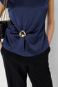 A person wearing a navy blue satin blouse with a drapé effect and a gold abstract brooch at the waist, black pants, and holding a black purse with a gold chain strap.
