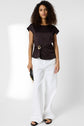 A woman with curly hair wears a black short-sleeve top with a bijou doré detail, white wide-leg pants, black sandals, and holds a small black clutch. She stands against a plain white background.
